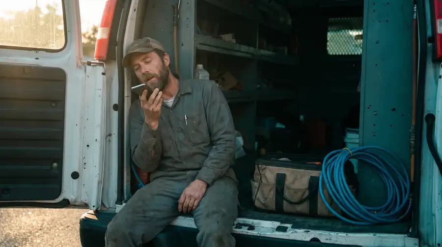 A mobile mechanic sitting on the open back of his work van, speaking a short voice memo into his phone.